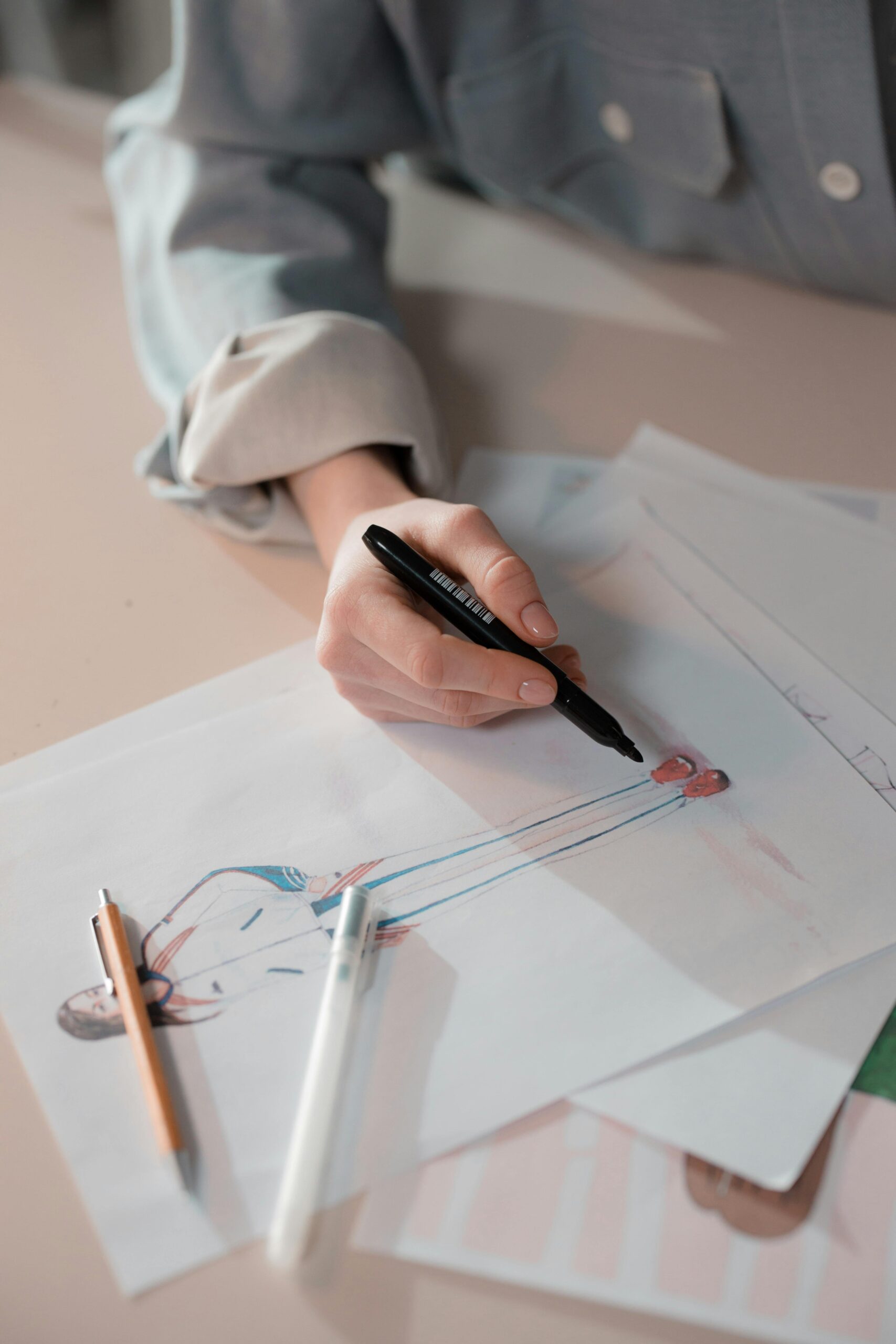 A hand sketching a fashion design with pens and papers on a table, showcasing creativity.